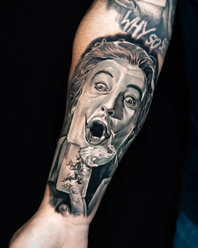 A forearm: a wide-eyed screaming face in white make-up, a playing card at the wrist